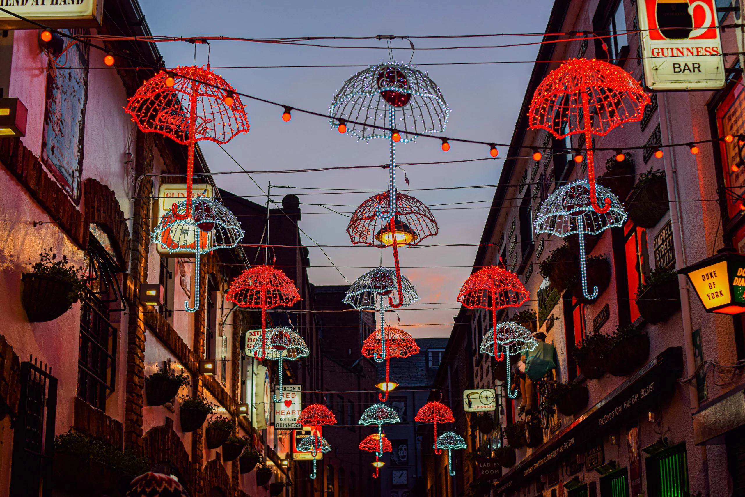 Nighttime view of Commercial Court in Belfast's Cathedral Quarter, featuring iconic hanging red and white light-up umbrellas between historic brick buildings.