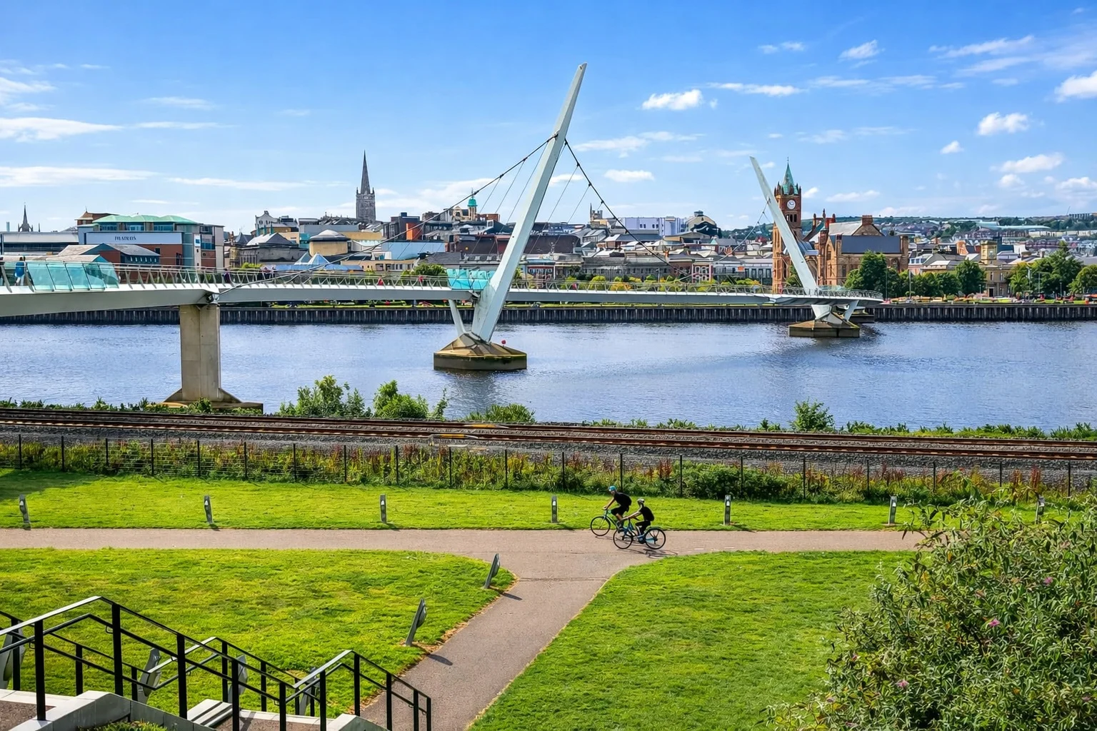 Cyclists riding along the River Foyle path with a view of the modern Peace Bridge and the historic Derry~Londonderry city skyline under a blue sky.