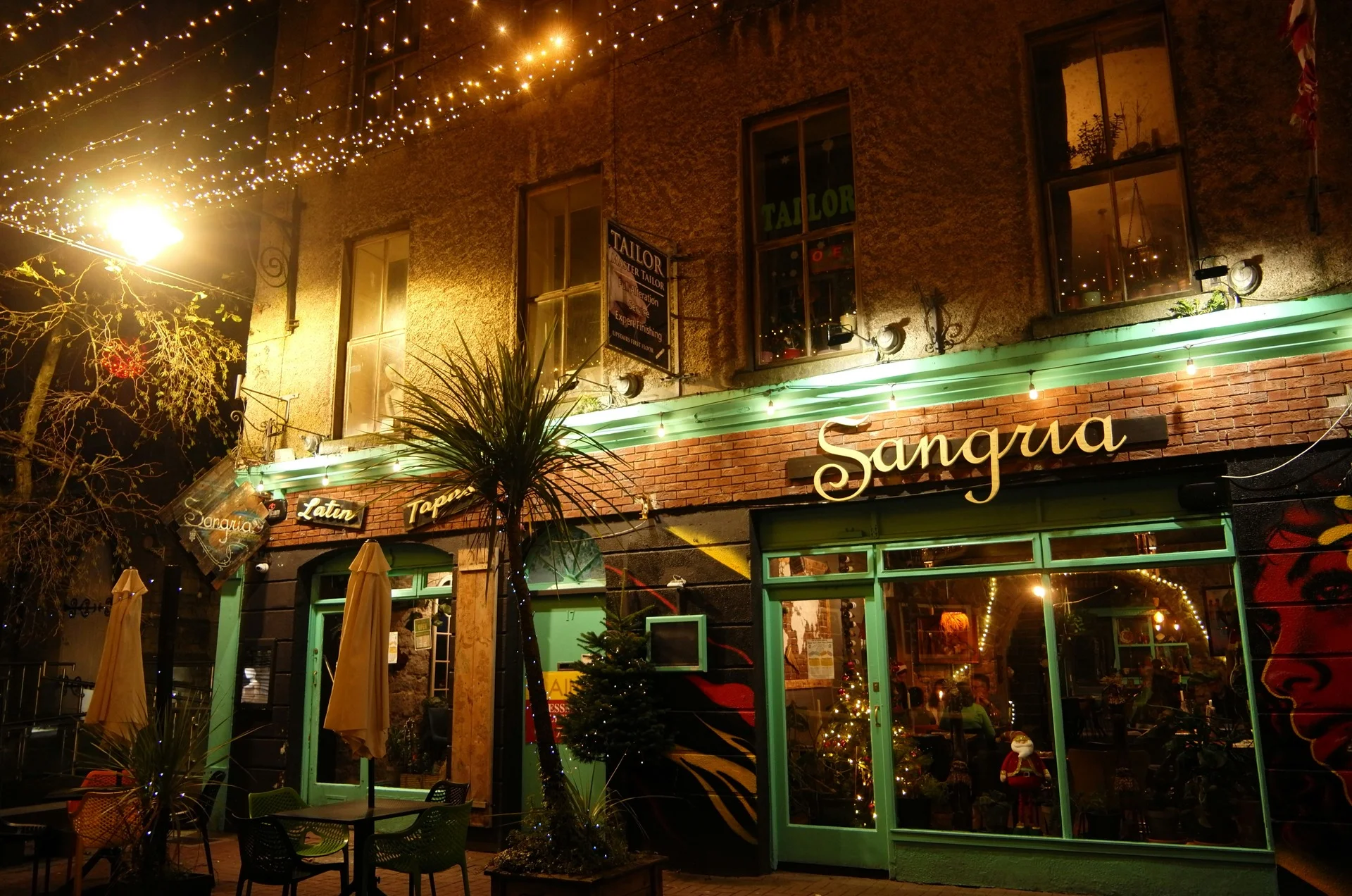 Nighttime exterior of Sangria tapas restaurant in Galway’s Latin Quarter, featuring glowing fairy lights, green window frames and outdoor seating.