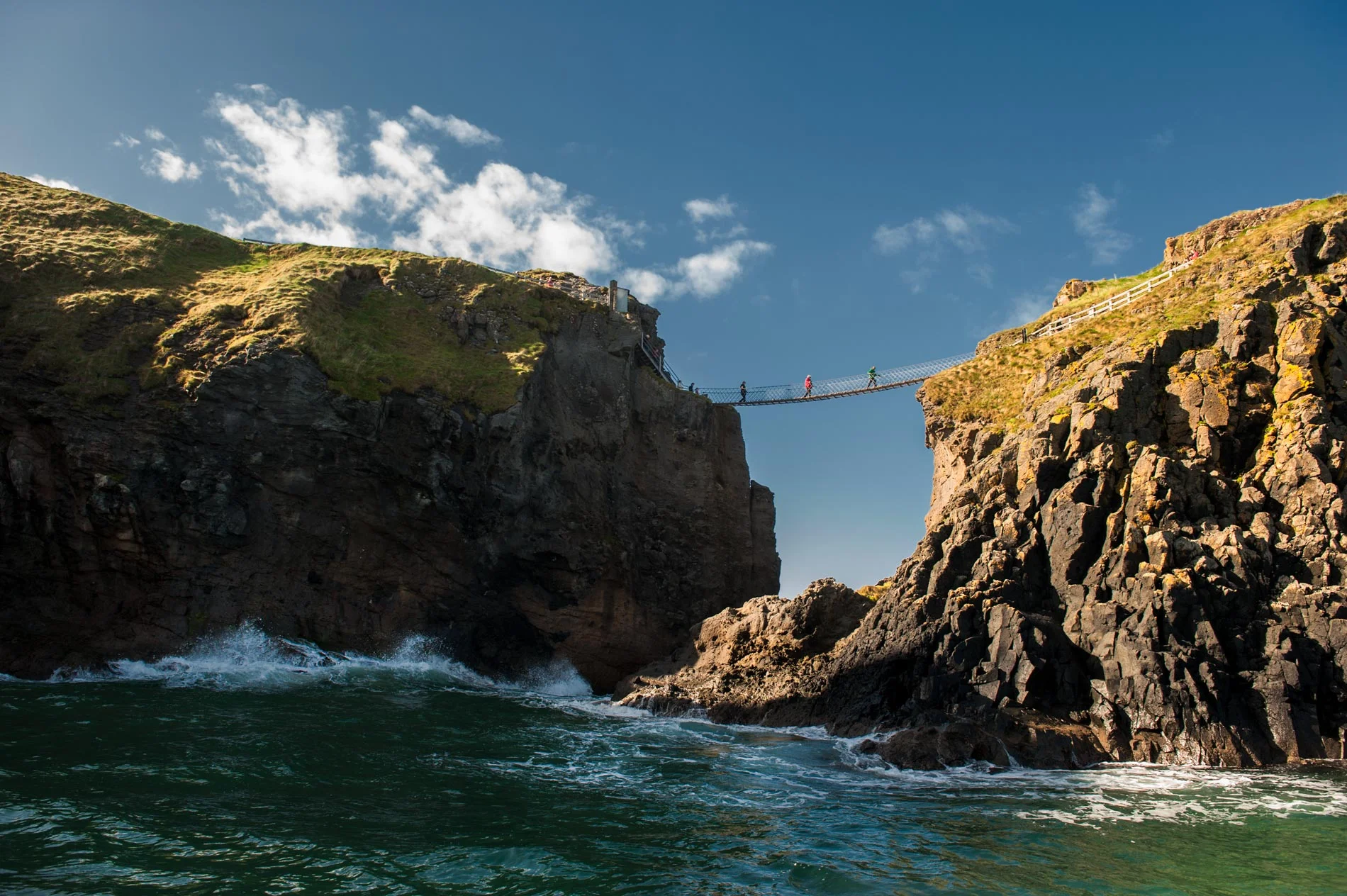People crossing the historic Carrick-a-Rede Rope Bridge suspended between rugged coastal cliffs above the turquoise Atlantic Ocean in Northern Ireland.