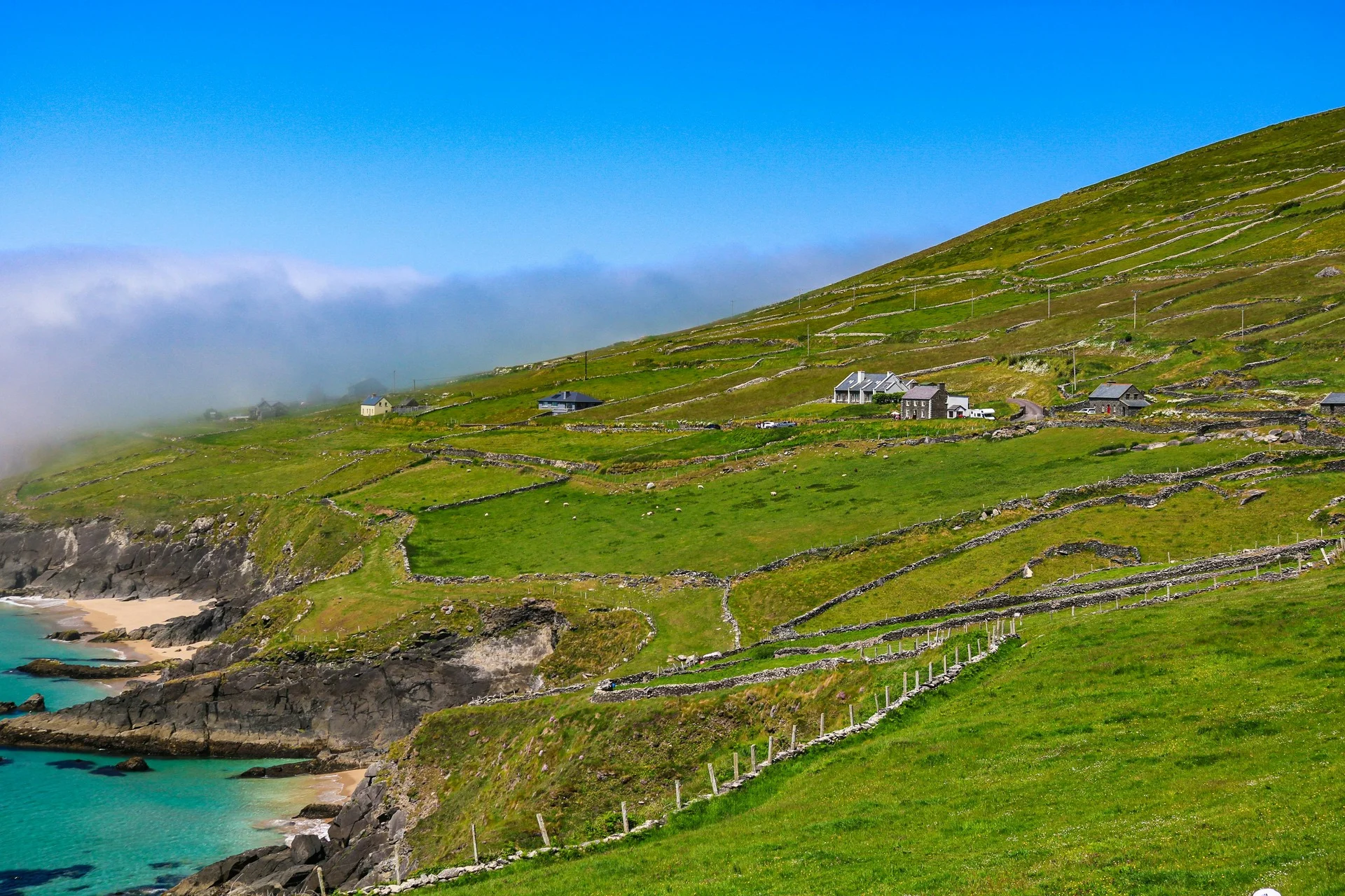 Lush green Irish hillside with traditional stone walls overlooking a turquoise bay and sandy beach under a bright blue sky.