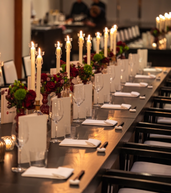 A long dark wood dining table set with white candles, floral centerpieces, and wine glasses in a dimly lit luxury venue.