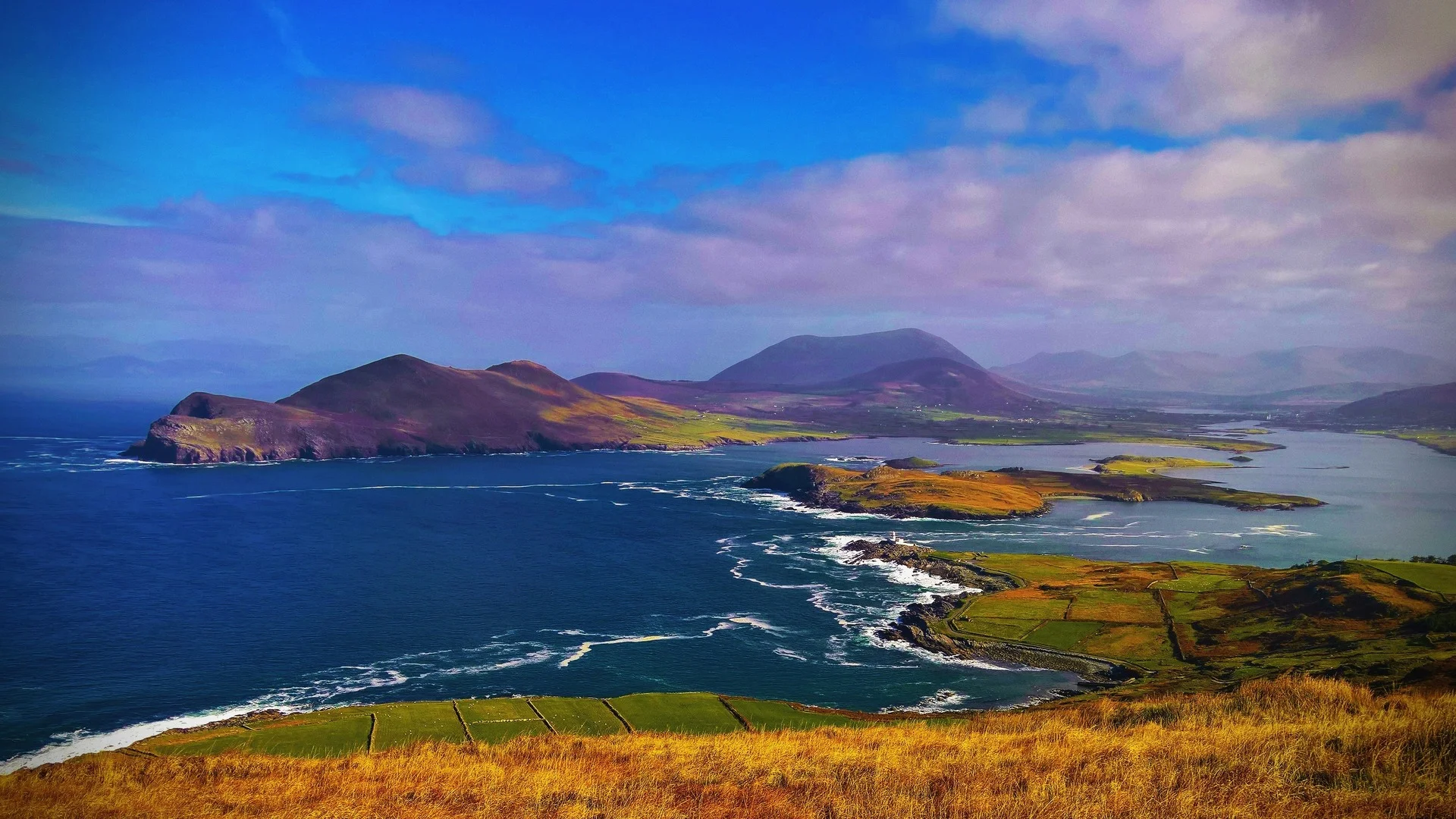 Panoramic view of the Ring of Kerry coastline featuring rolling green fields, rugged islands, and deep blue Atlantic waters under a cloudy sky.