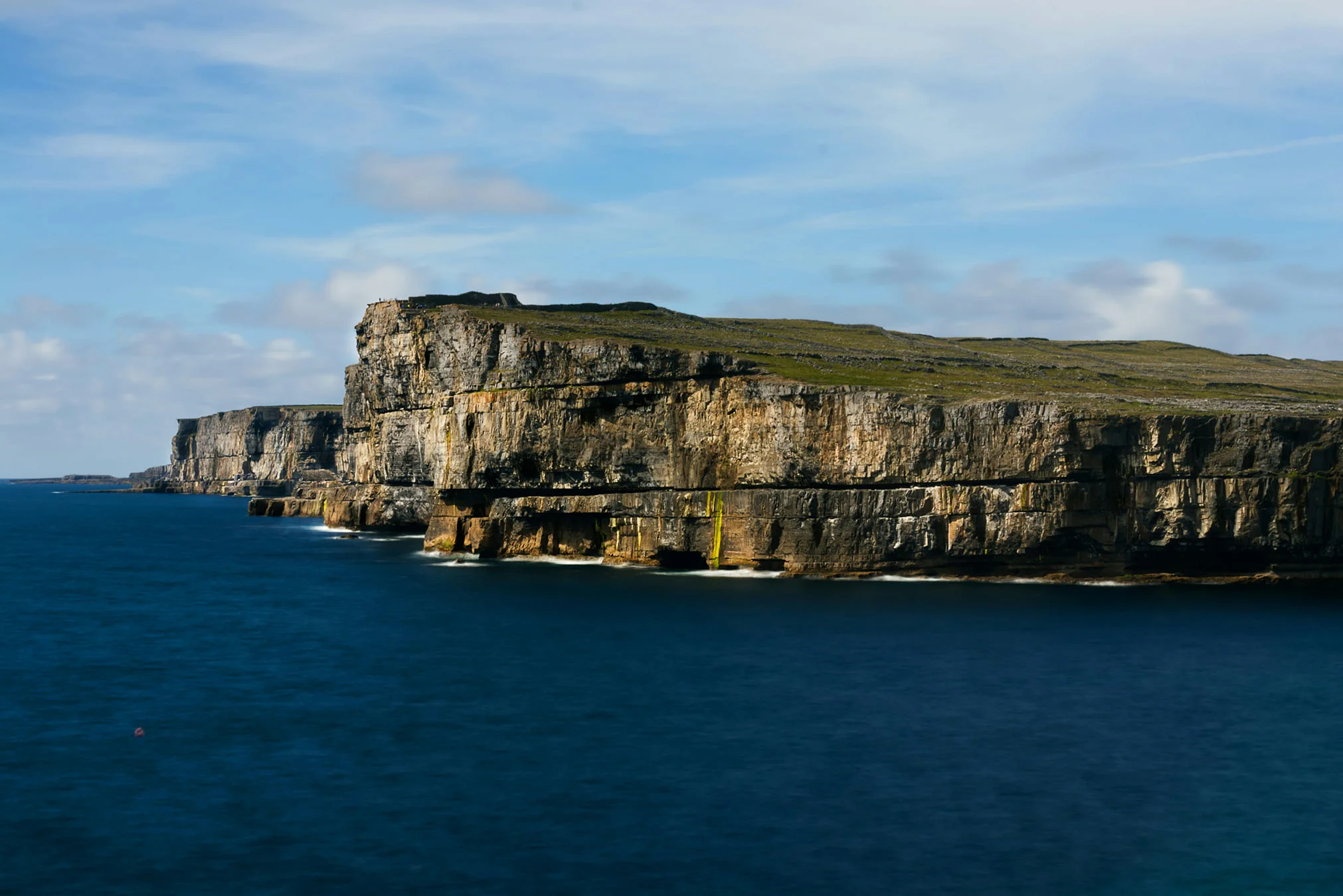 Wide-angle view of the sheer limestone sea cliffs of Inis Mór, Aran Islands, under a bright blue sky with calm ocean water.