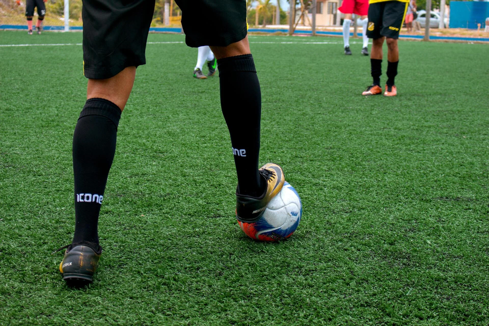 A close-up of a soccer player's legs in black socks and cleats with one foot resting on a ball on a green artificial turf field.