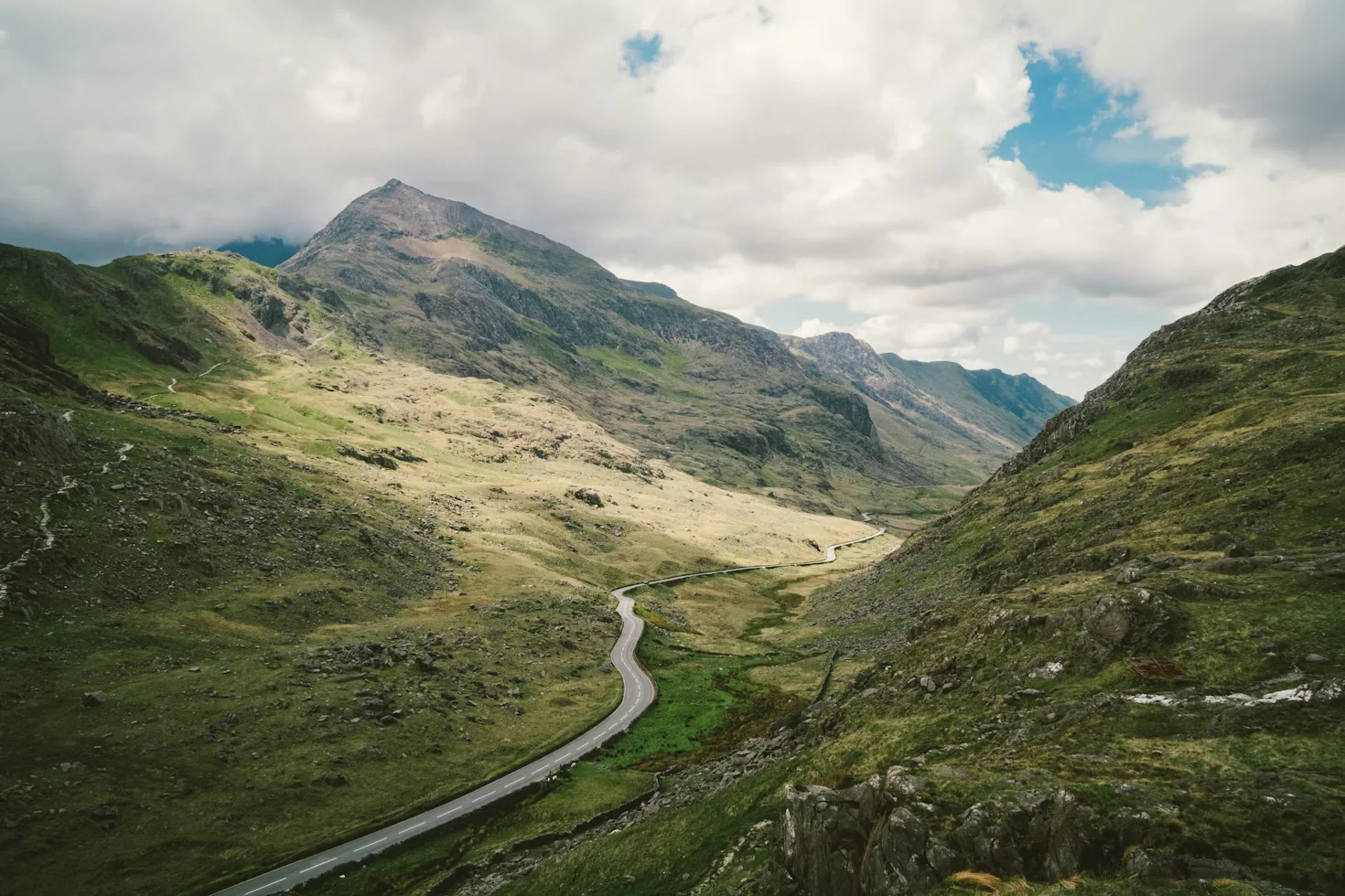 A winding asphalt road cutting through the rugged, grassy mountains of Snowdonia National Park in Wales under a cloudy blue sky.