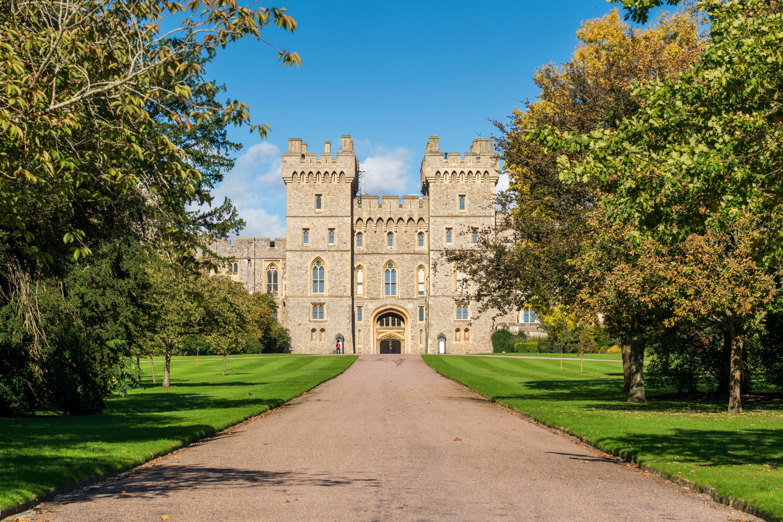Tree-lined pathway leading toward a historic English royal residence under blue sky